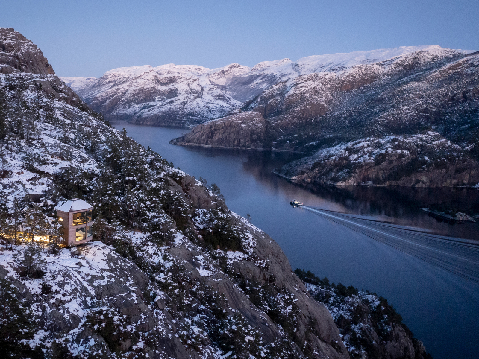Oversiktbilde av Lysefjorden med en båt på vannet og en hytte som lyser opp i fjellveggen. Foto.