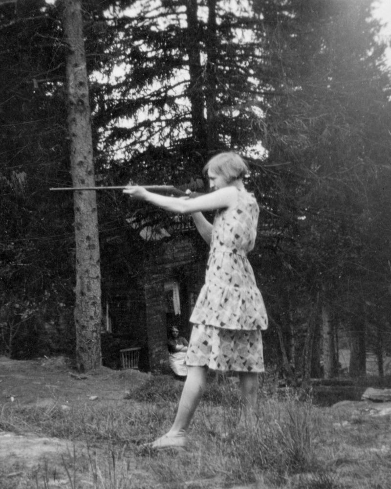 Kvinne i kjole står og sikter med et skytevåpen, svart hvitt. Foto.