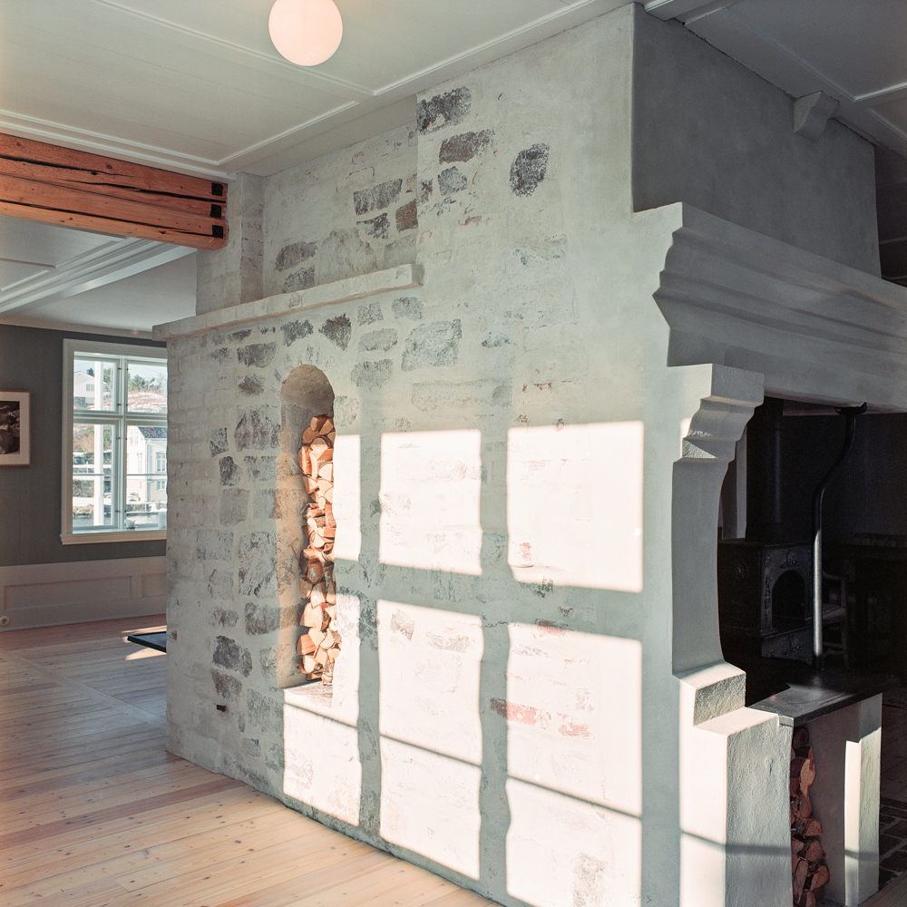 Interiør foto, hvit pusset teglvegg midt i rommet, åpning i vegg fylt av stablet ved. 