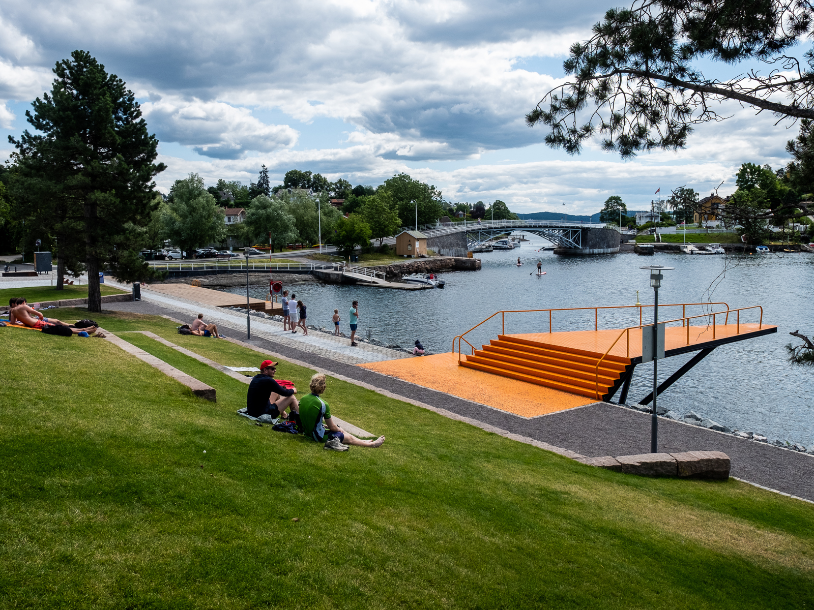 Grønn park med mennesker, oransje stupebrett ved vannkanten. Foto.