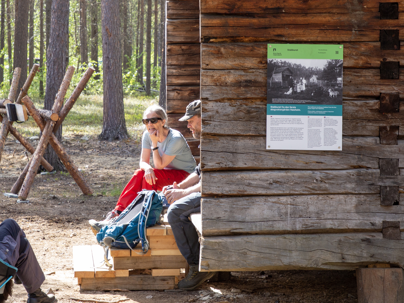 Tømret hytte, to mennesker sitter i inngangsparti, skog i bakgrunnen. Foto.