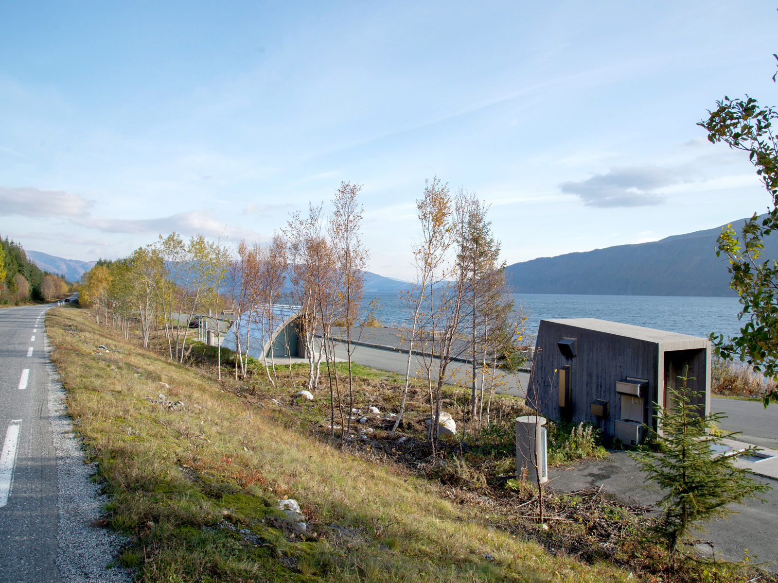 Fotografi tatt fra vei ned på rasteplass med to mindre bygg, utsikt til fjell og vann.