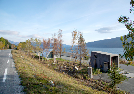 Fotografi tatt fra vei ned på rasteplass med to mindre bygg, utsikt til fjell og vann.