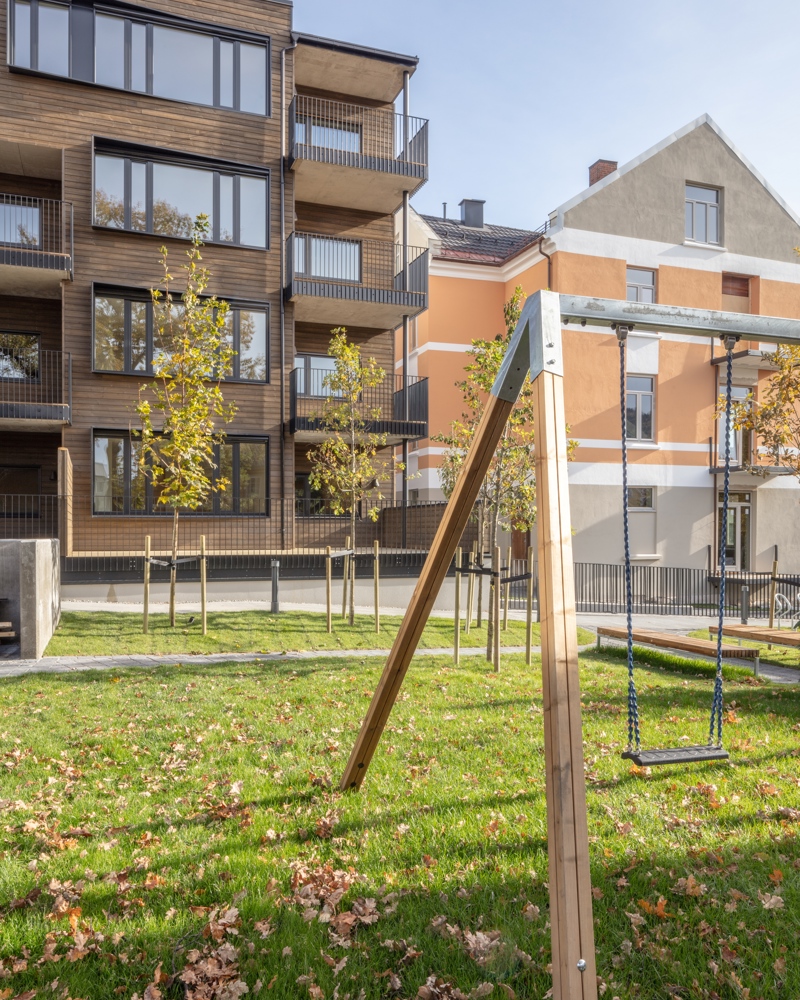 Bakgård, huske stativ i front, grønn plen, i bakgrunnen boligblokk i treverk møter klassisk bygårdsbygg. Foto.